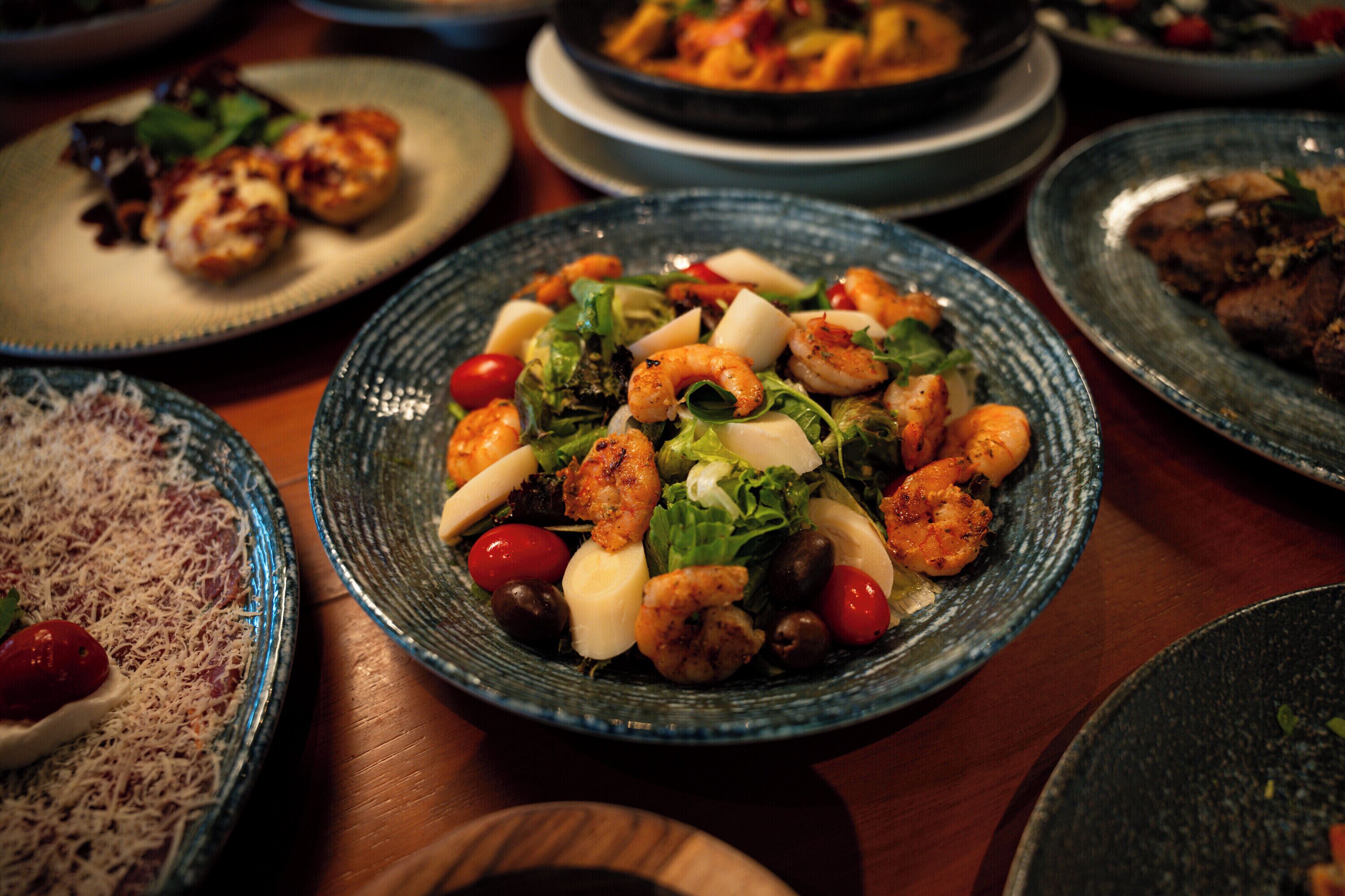 Variedad de platillos en una mesa, incluyendo una ensalada de camarones con verduras y queso.