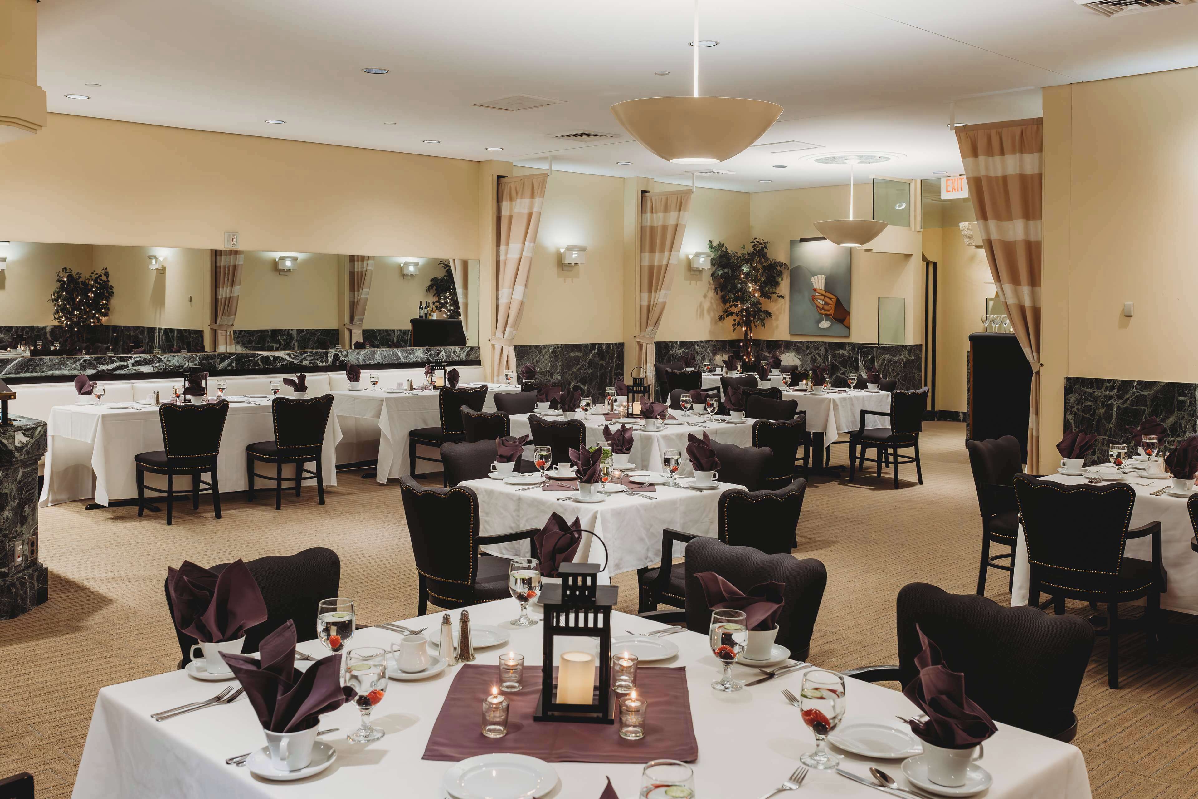 A hotel dining area with multiple tables set for dining, each with white tablecloths, dark chairs, and candles.