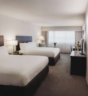 A hotel room with two beds, a desk, a lamp, a vase with flowers, and a large window with curtains.