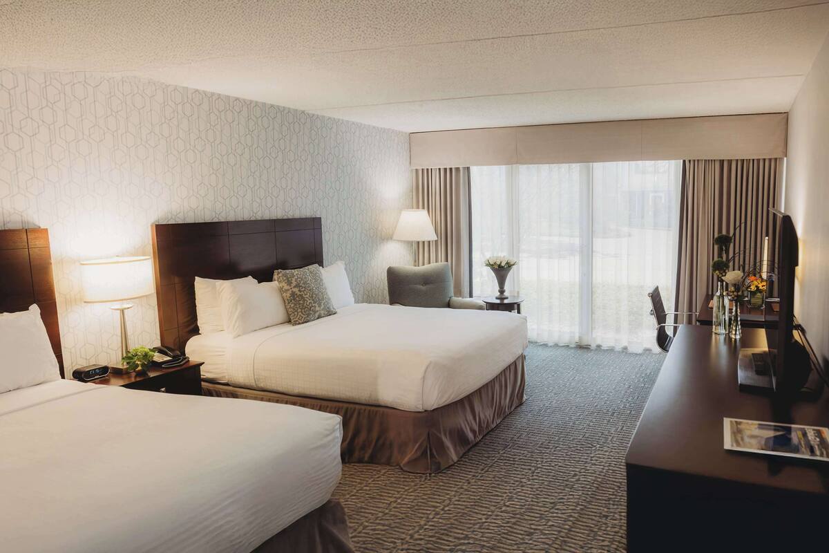 A hotel room with two beds, a desk, a lamp, a clock, and a window with curtains.