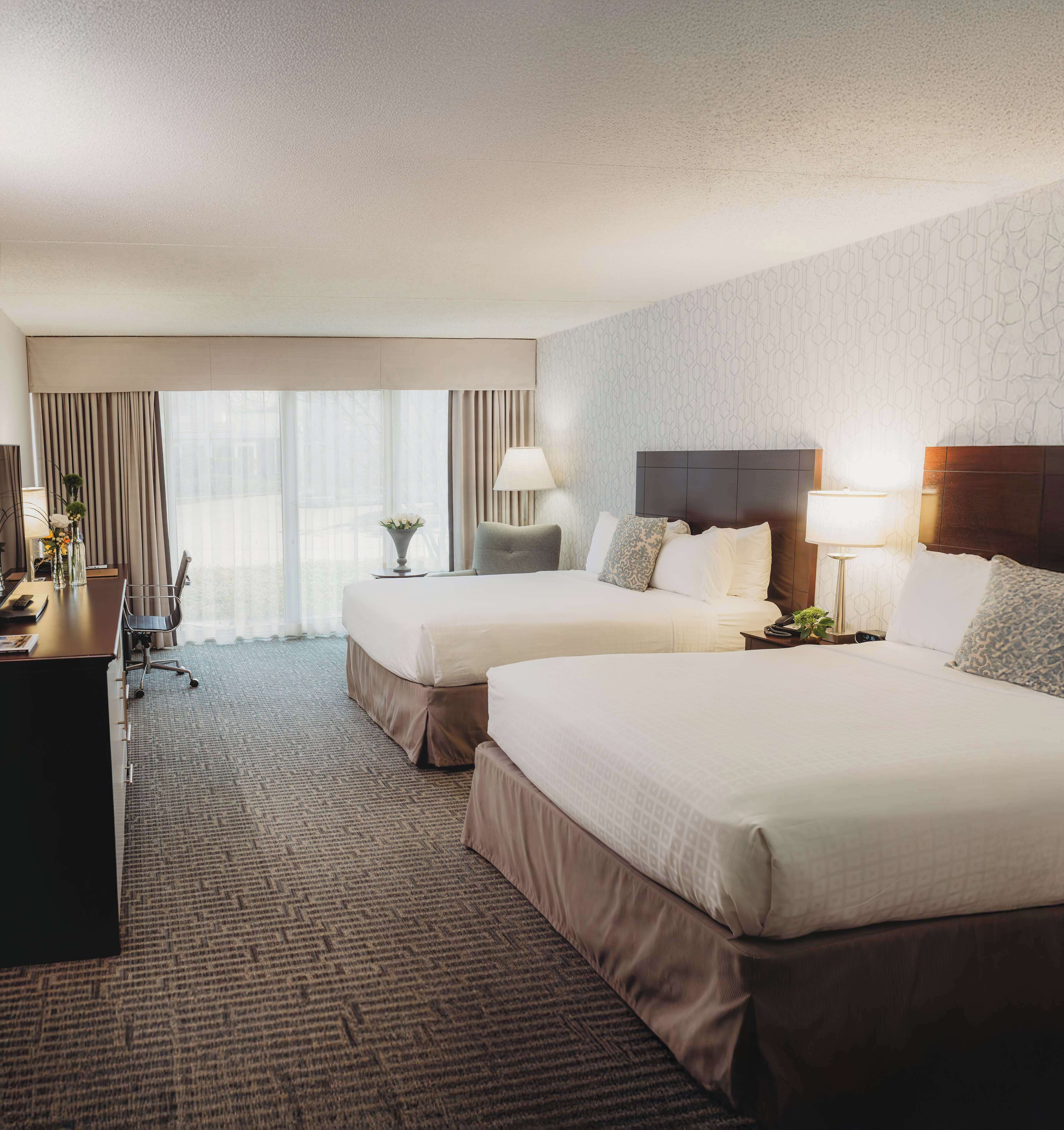 A hotel room with two beds, a desk, a chair, a lamp, and a view of the outdoors.