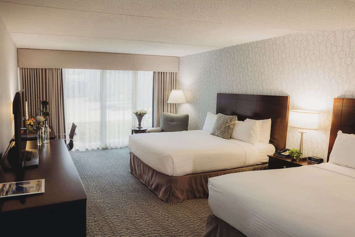 A hotel room with two beds, a desk, a chair, a lamp, and a view of the outdoors.