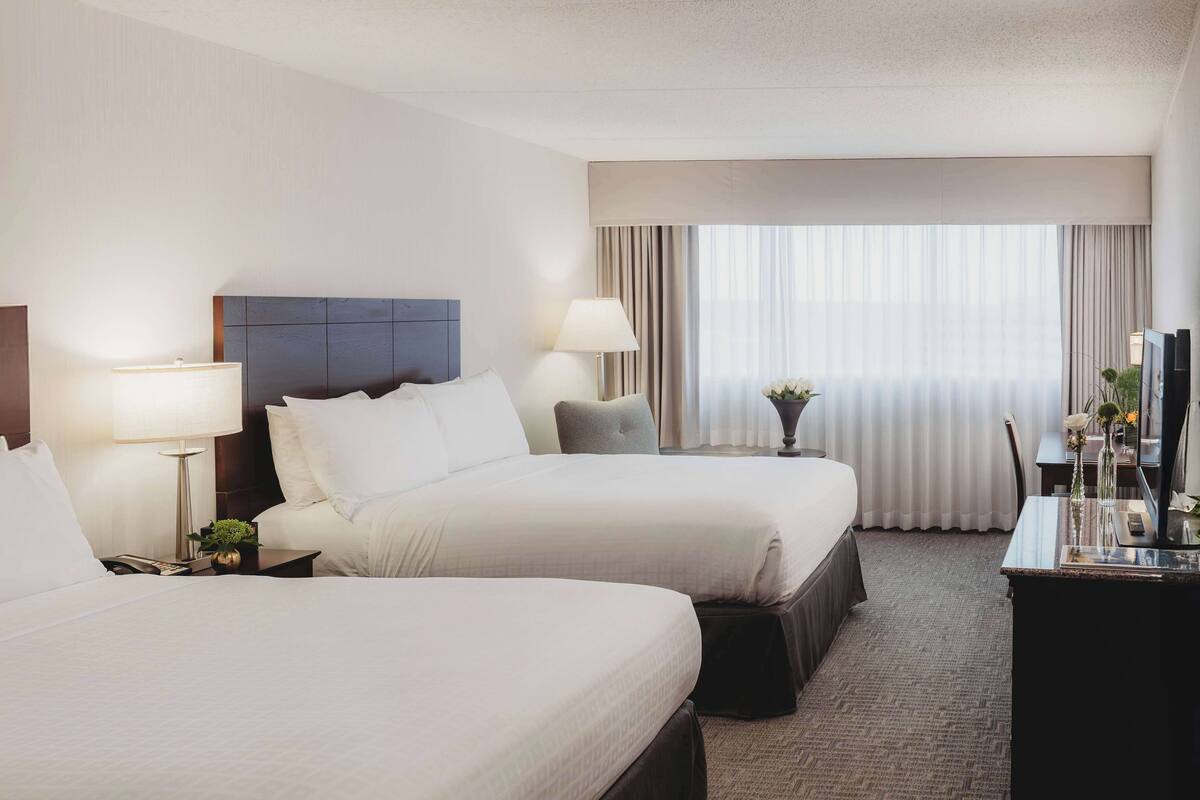 A hotel room with two beds, a desk, a lamp, a vase with flowers, and a window with curtains.