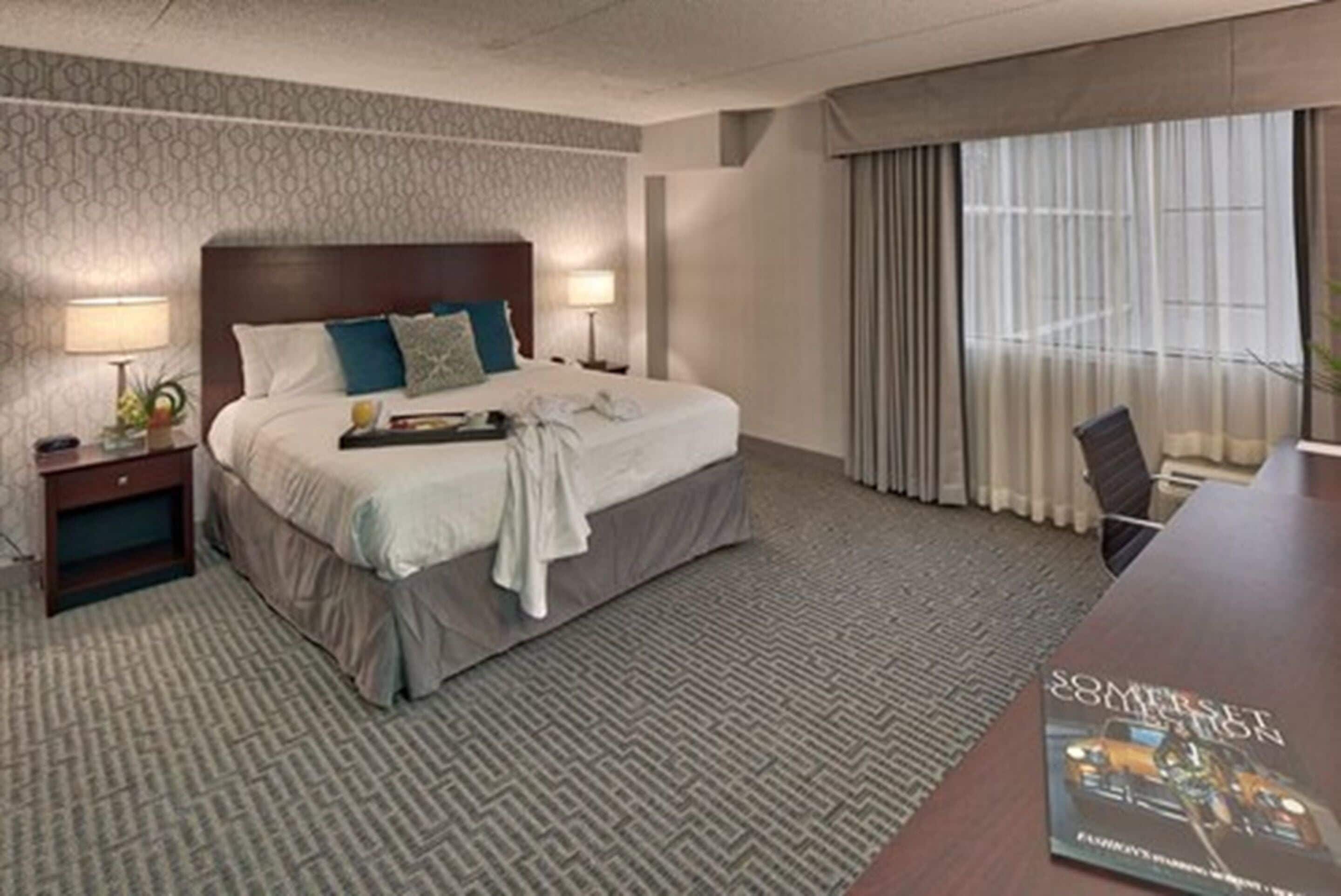 Een hotelkamer met een groot bed, twee nachtkastjes met lampen, een bureau met stoel, en een groot raam met gordijnen.