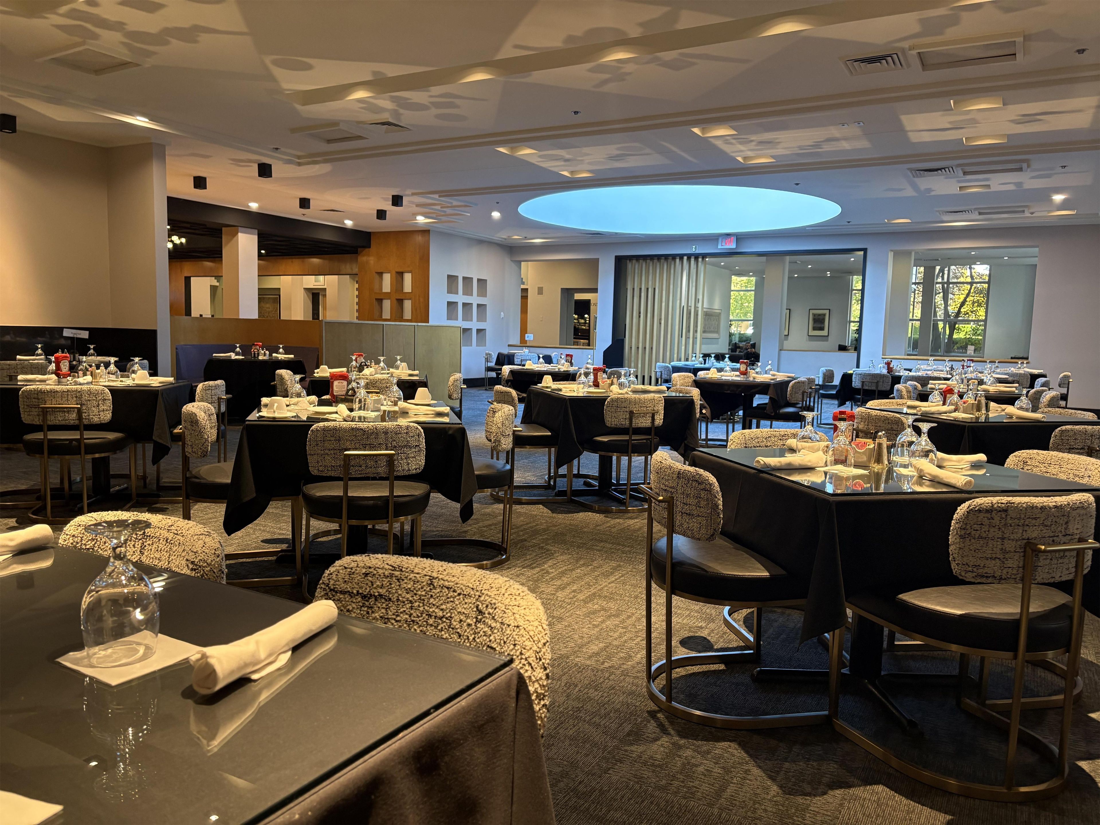 A hotel dining area with multiple tables set for service, a central skylight, and a view of the outdoors.