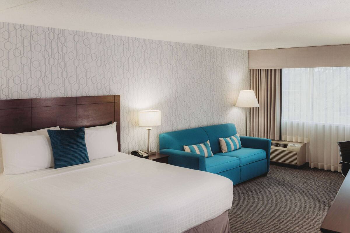 A hotel room with a large bed, a blue sofa, a nightstand with a lamp, and a window with curtains.