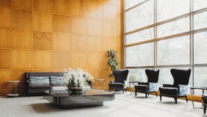 A modern living room with a wooden paneled wall, a large window, a sofa, armchairs, and a coffee table with a floral arrangement. - Somerset Inn, WorldHotels Distinctive (Troy)