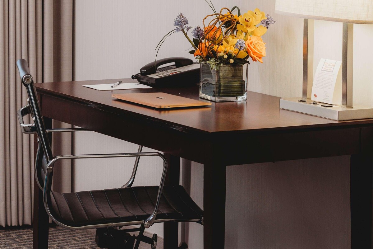 A hotel room with a wooden desk, a black office chair, a vase with flowers, a telephone, a notepad, a lamp, and a sign on the wall.