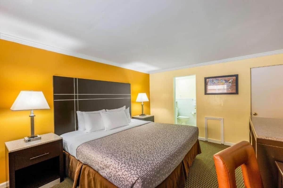 Habitación de hotel con cama, dos lámparas de noche, mesita de noche, una silla y puerta que da a un baño.