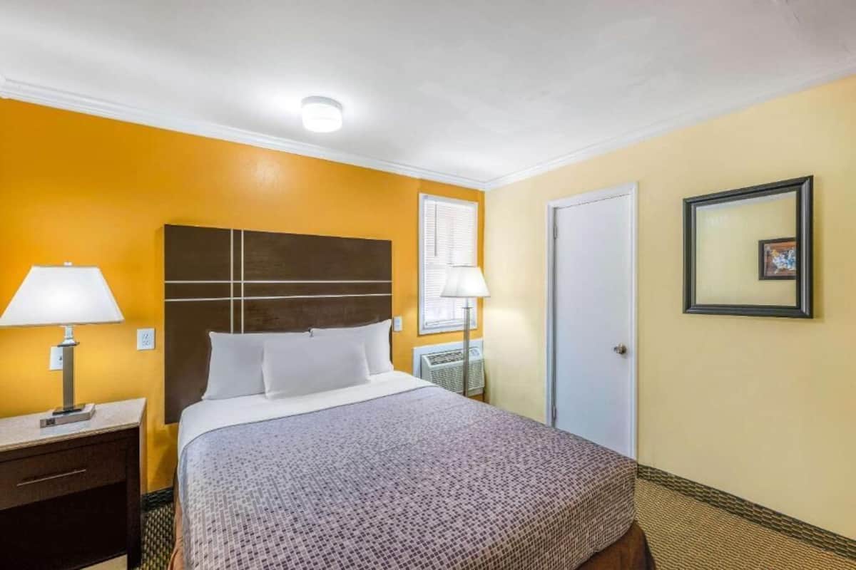Habitación de hotel con cama, lámparas de noche, mesita de noche, una puerta y un cuadro en la pared.