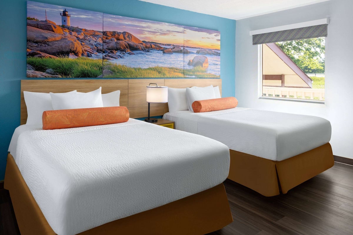 A hotel room with two beds, a large painting of a coastal scene, a nightstand with a lamp, and a window with a view of the outdoors.