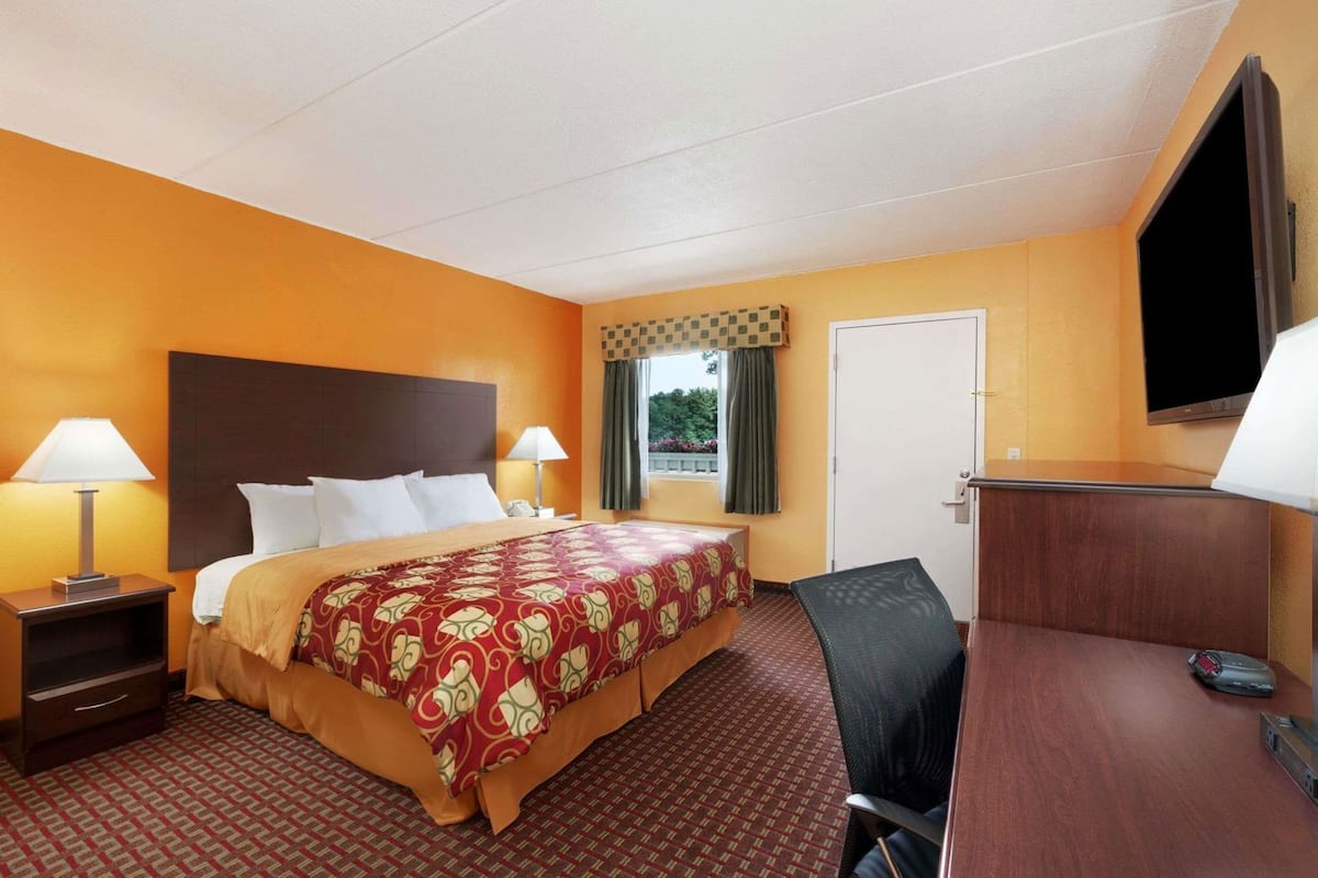 A hotel room with a bed, two bedside tables with lamps, a desk with a chair, a TV, and a window with curtains.