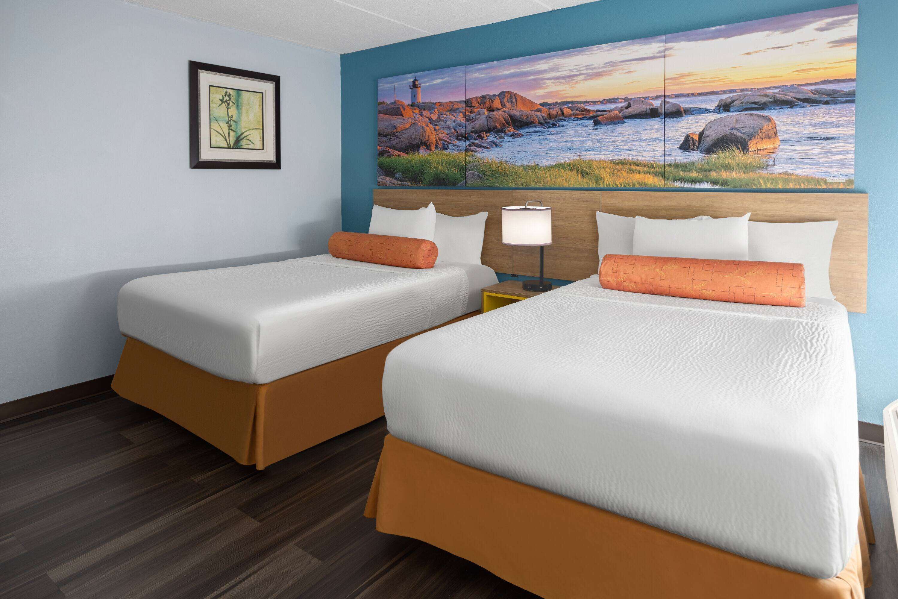 A hotel room with two beds, a nightstand, a lamp, a framed picture, and a large painting of a coastal scene.