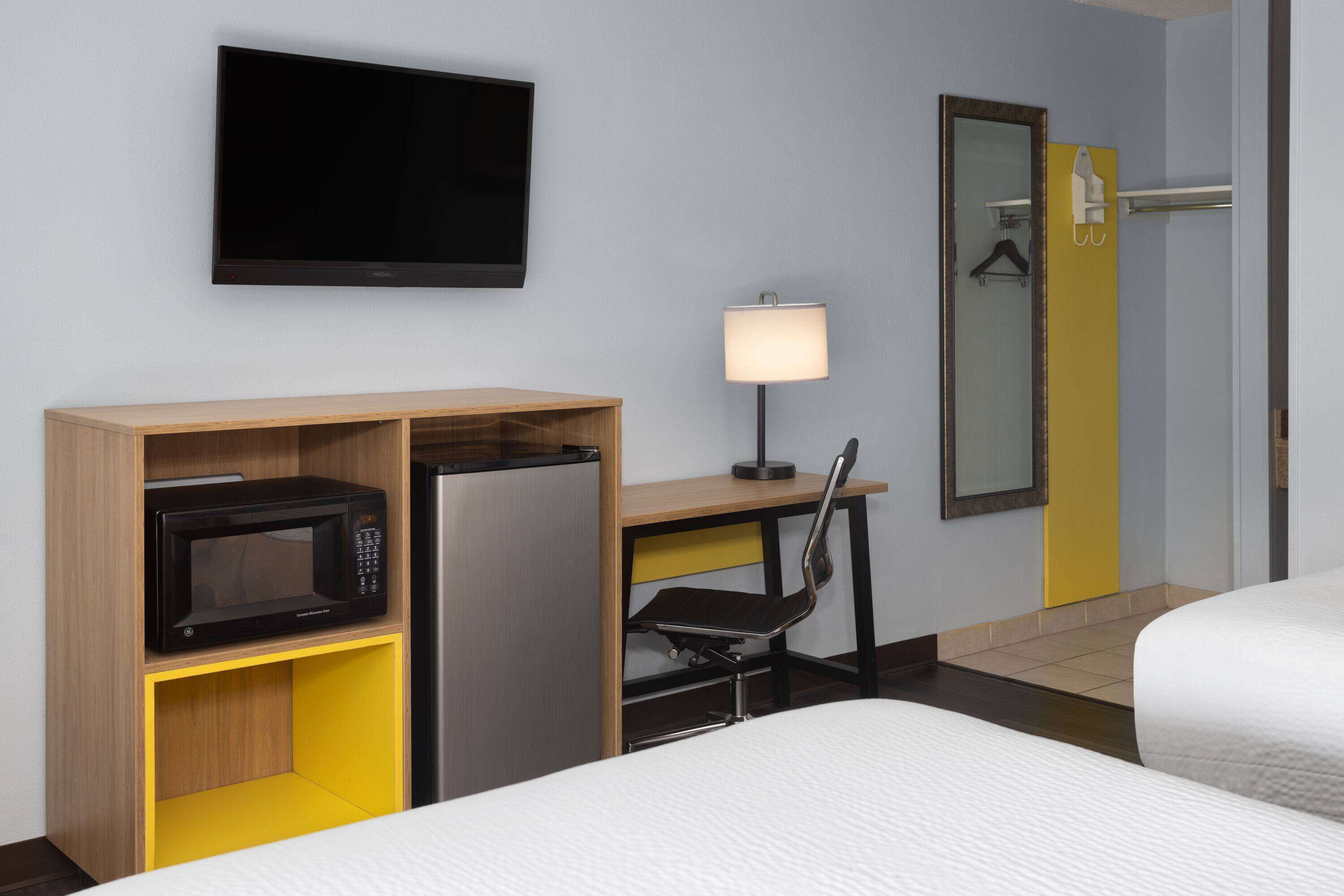 A hotel room with a bed, a desk with a microwave, a chair, a TV, and a bathroom door.