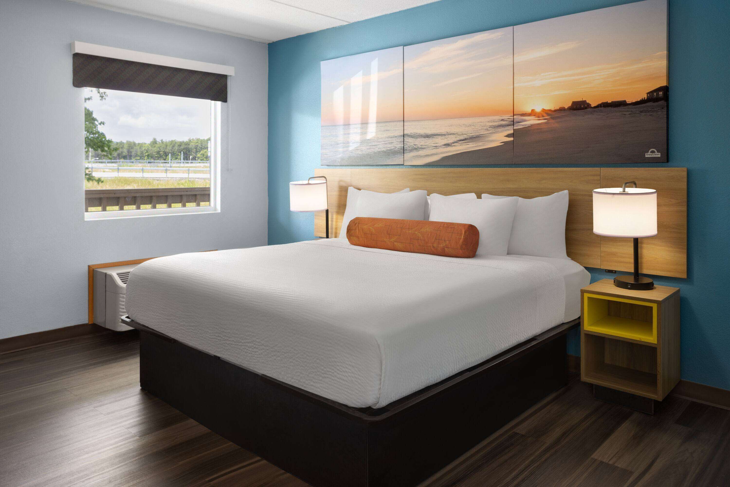A hotel room with a large bed, a nightstand with a lamp, a window with a view of the beach, and a sunset painting on the wall.