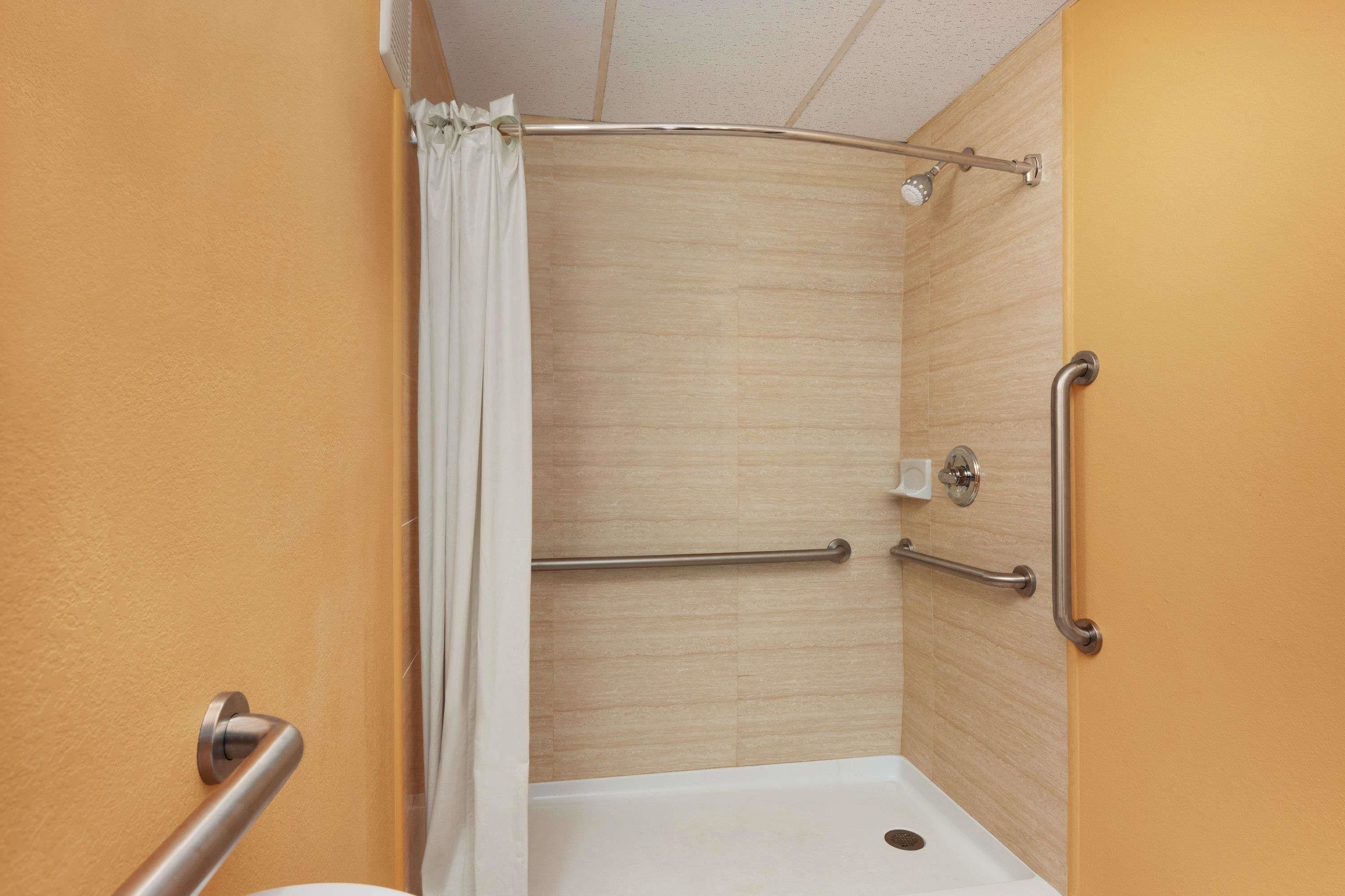 Een badkamer met een douche, een witte badkuip en een toilet. Er zijn handgrepen en een veiligheidsbeugel aanwezig.
