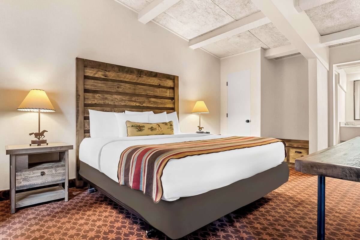 A bedroom with a wooden headboard, a bed with white linens, two bedside tables with lamps, and a patterned carpet.
