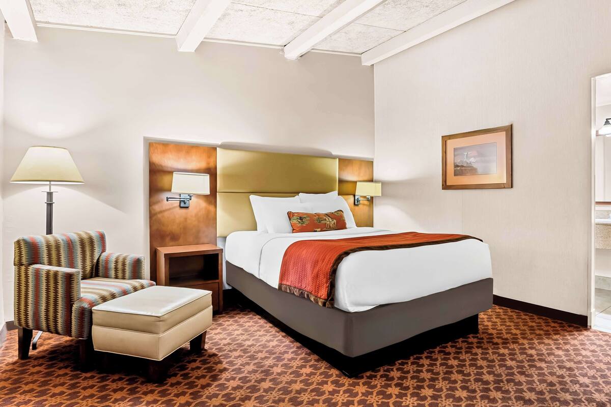 A hotel room with a bed, a chair, an ottoman, a nightstand, and a lamp.