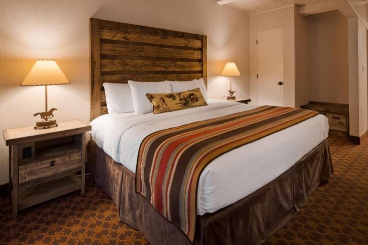 A hotel room with a large bed, two bedside lamps, a wooden headboard, a patterned carpet, and a bathroom with a mirror and sink.