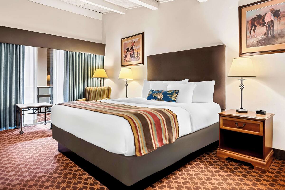 A hotel room with a large bed, two bedside tables with lamps, a small table, and a chair. There are two paintings on the walls.