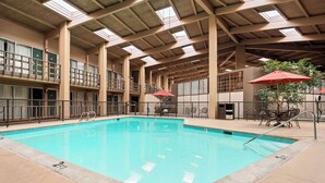 Una piscina interior con techo alto y tragaluces, rodeada por un balcón con barandillas y una zona de estar con sombrillas.
