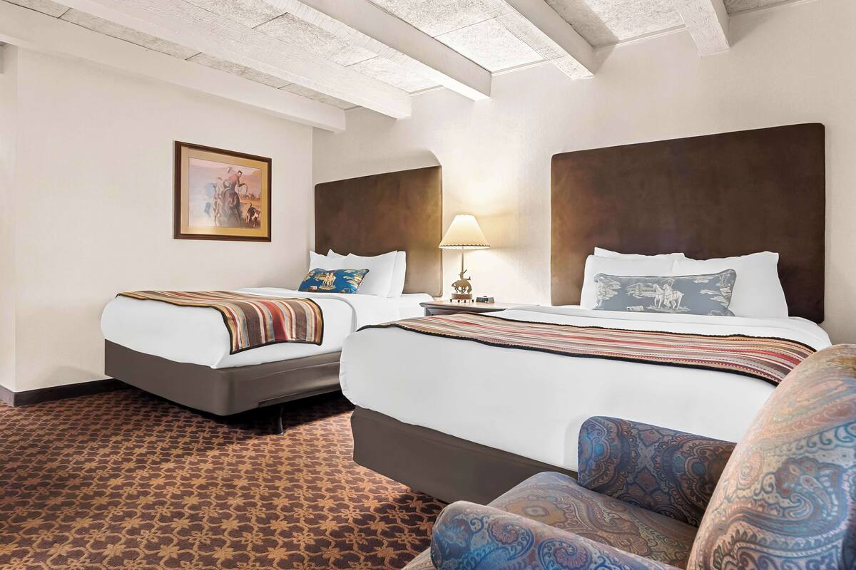 A hotel room with two beds, a patterned carpet, a framed picture on the wall, and a lamp on the nightstand.