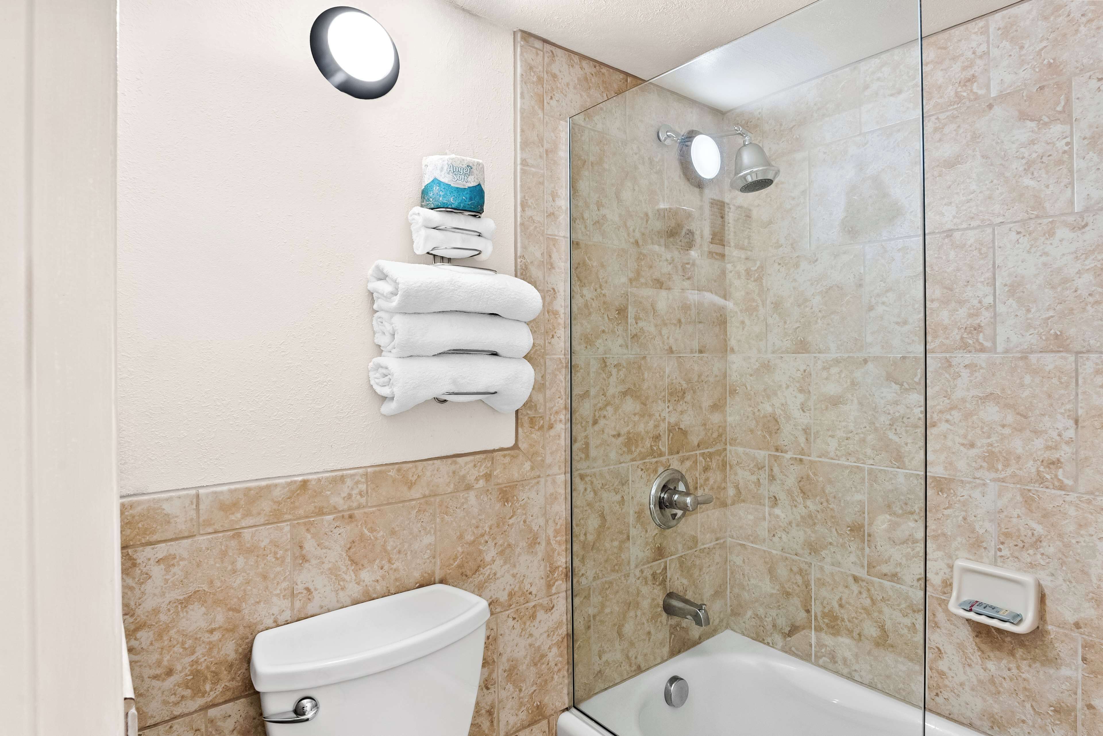 Un baño con un inodoro, una pila de toallas blancas, una ducha con cerramiento de vidrio y un jabonera.