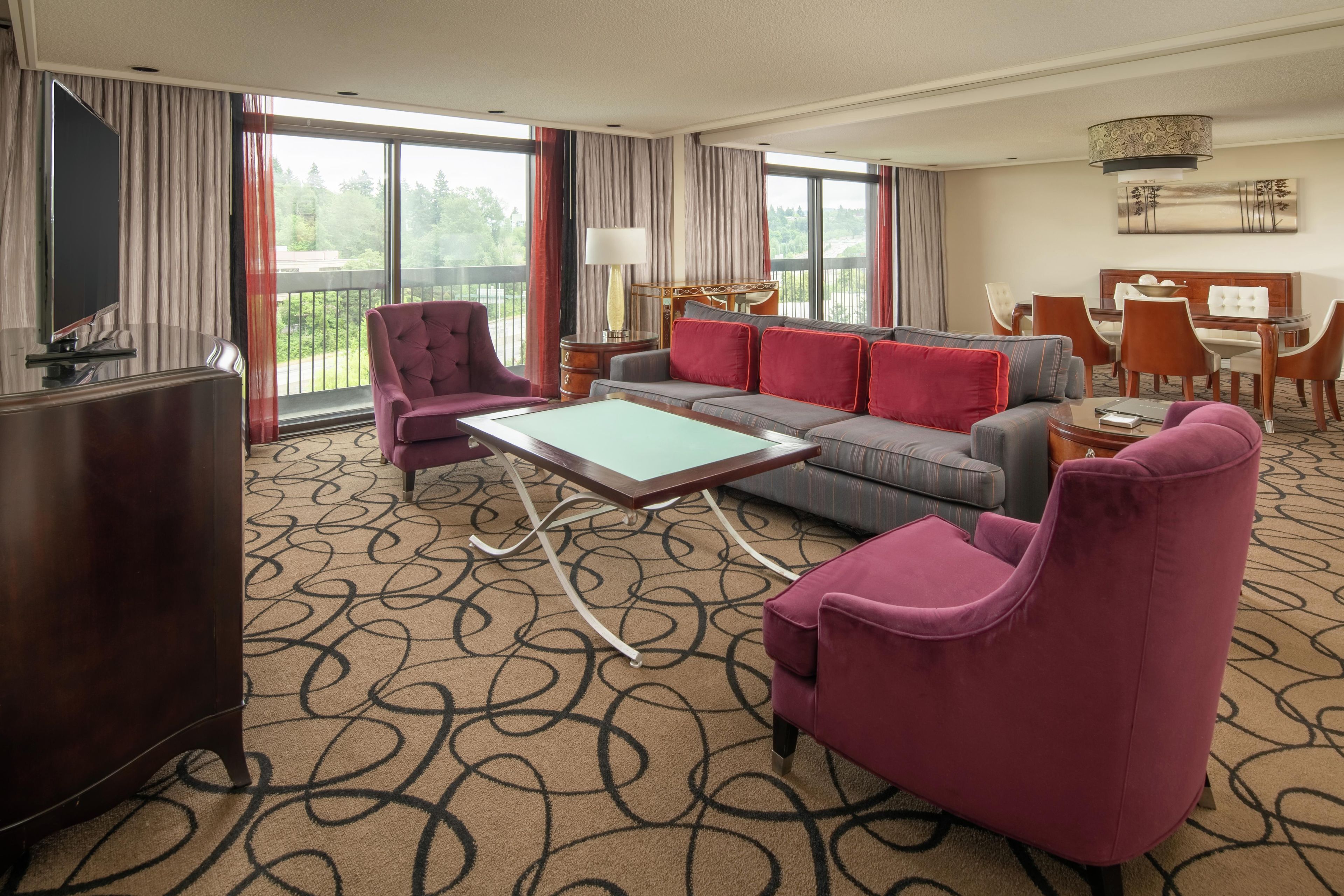 A hotel room with a sofa, armchairs, a coffee table, a TV, and a dining area with chairs and a table.