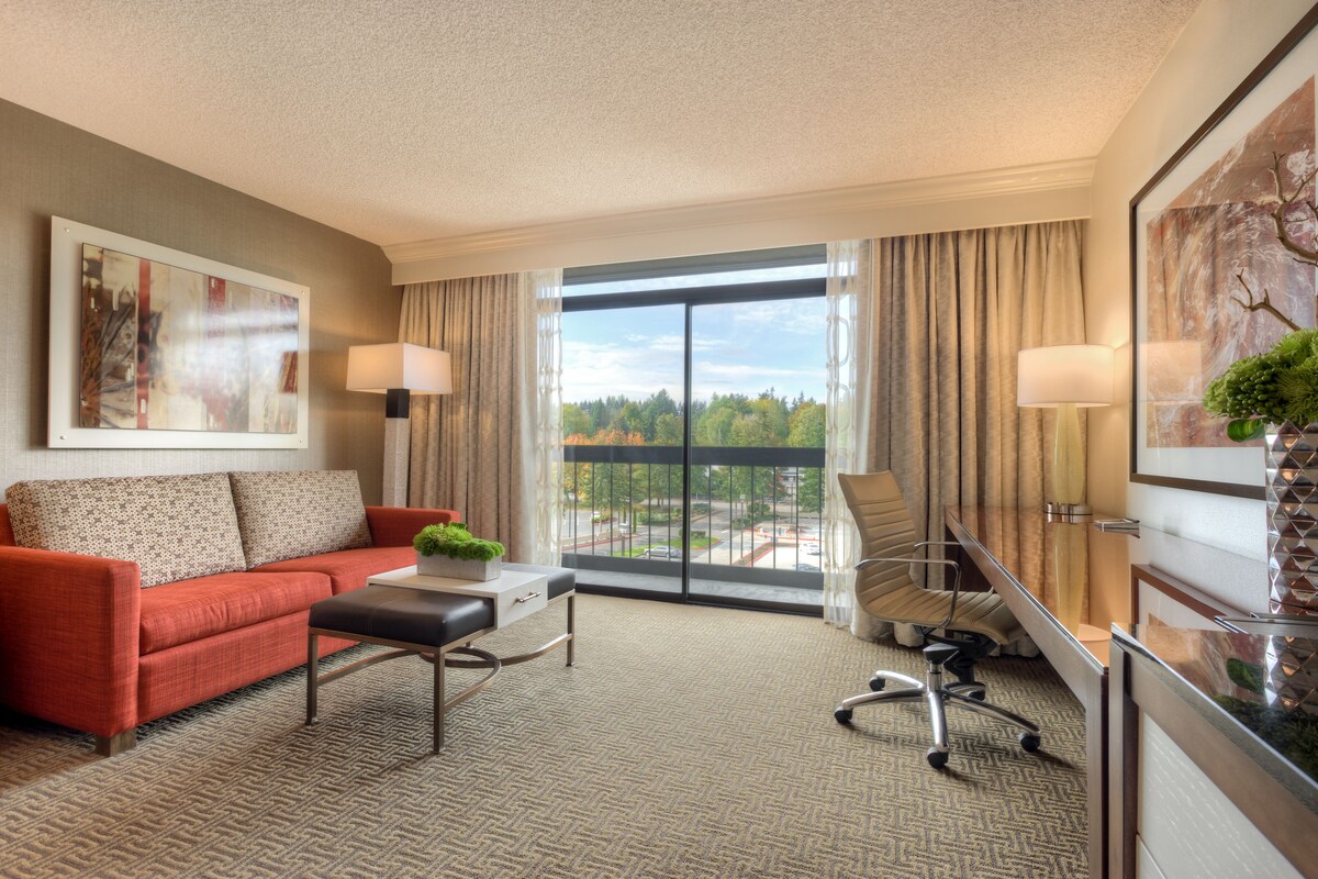 A hotel room with a red sofa, a desk, a chair, and a large window with a view of trees and buildings.