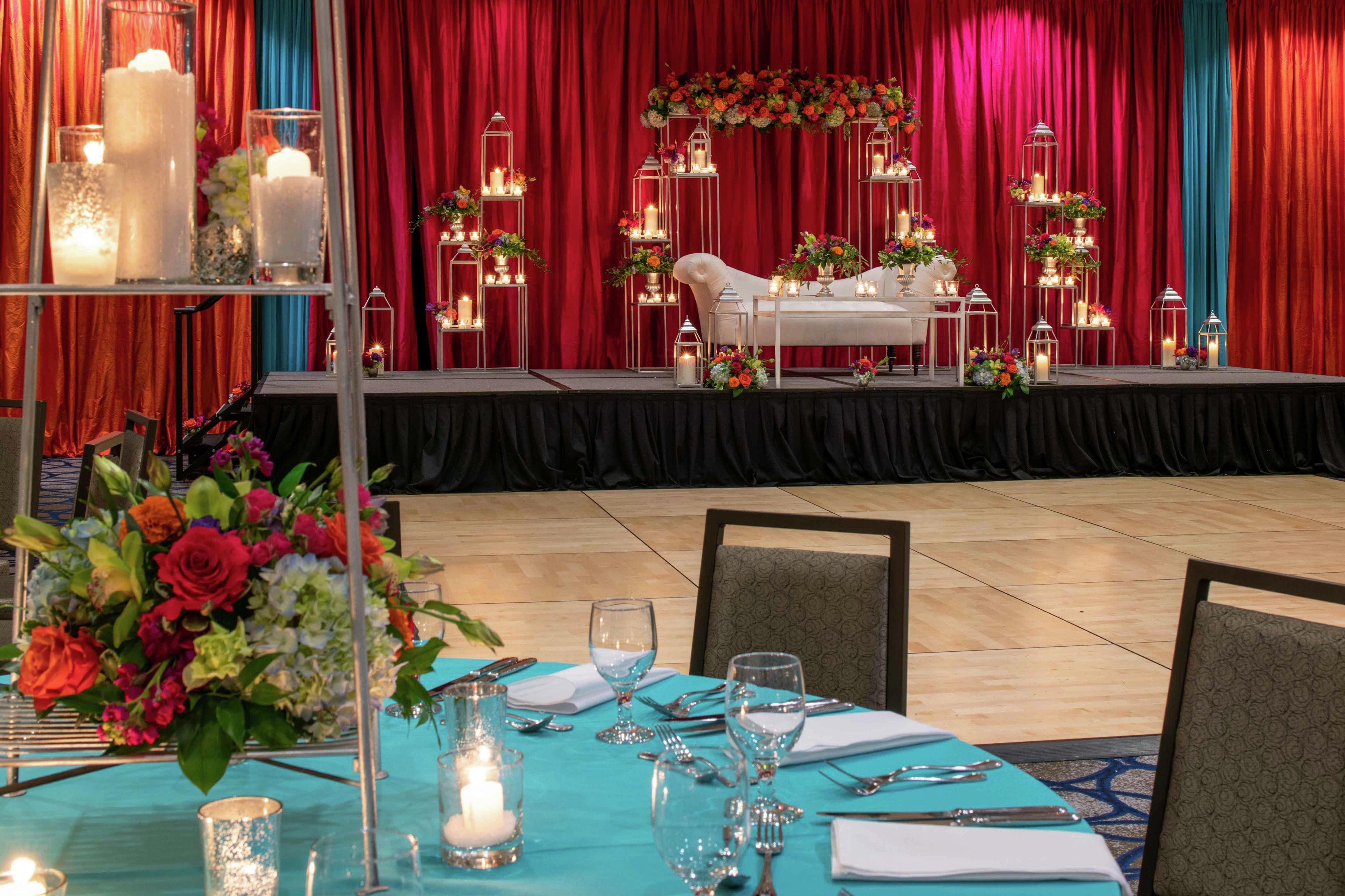 Un montaje formal para el comedor con una mesa dispuesta para cuatro personas, un escenario con un arreglo floral y cortinas rojas al fondo.