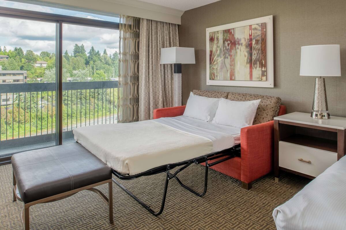A hotel room with a bed, a bench, a nightstand, a lamp, and a painting on the wall.
