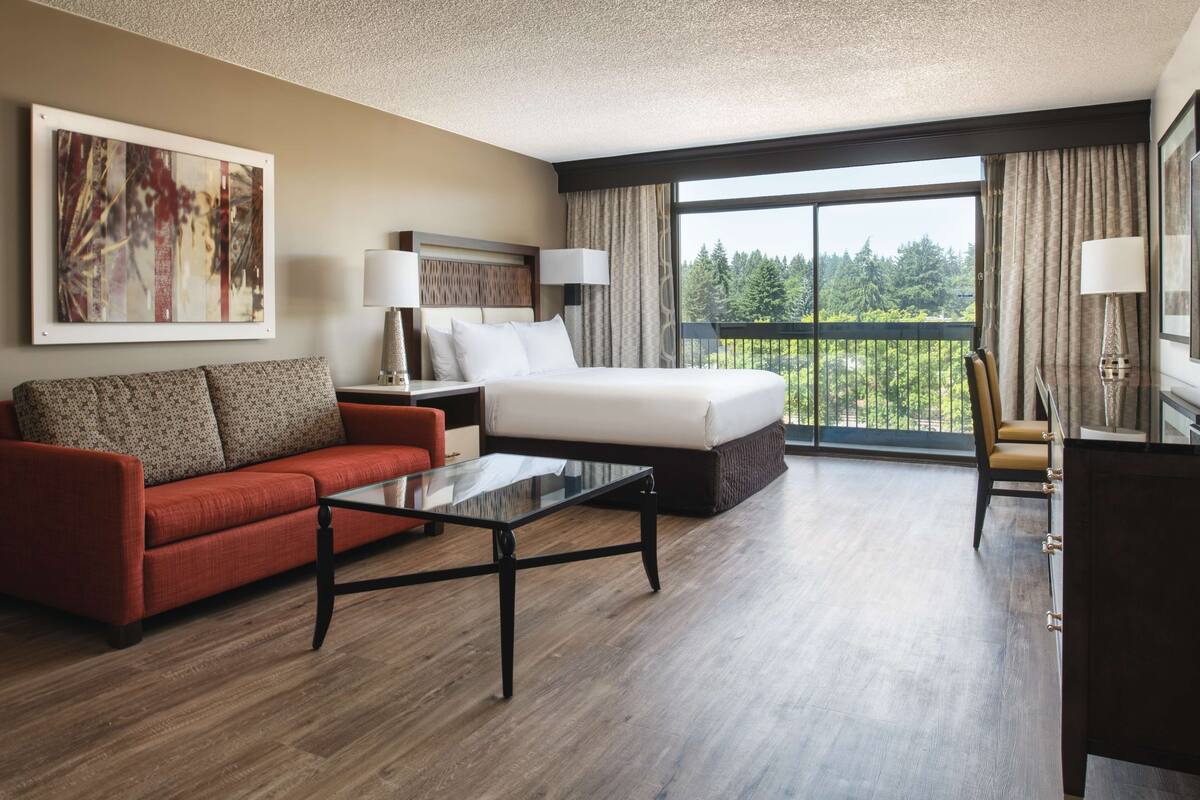A hotel room with a bed, a red sofa, a glass coffee table, a desk, and a view of greenery through large windows.