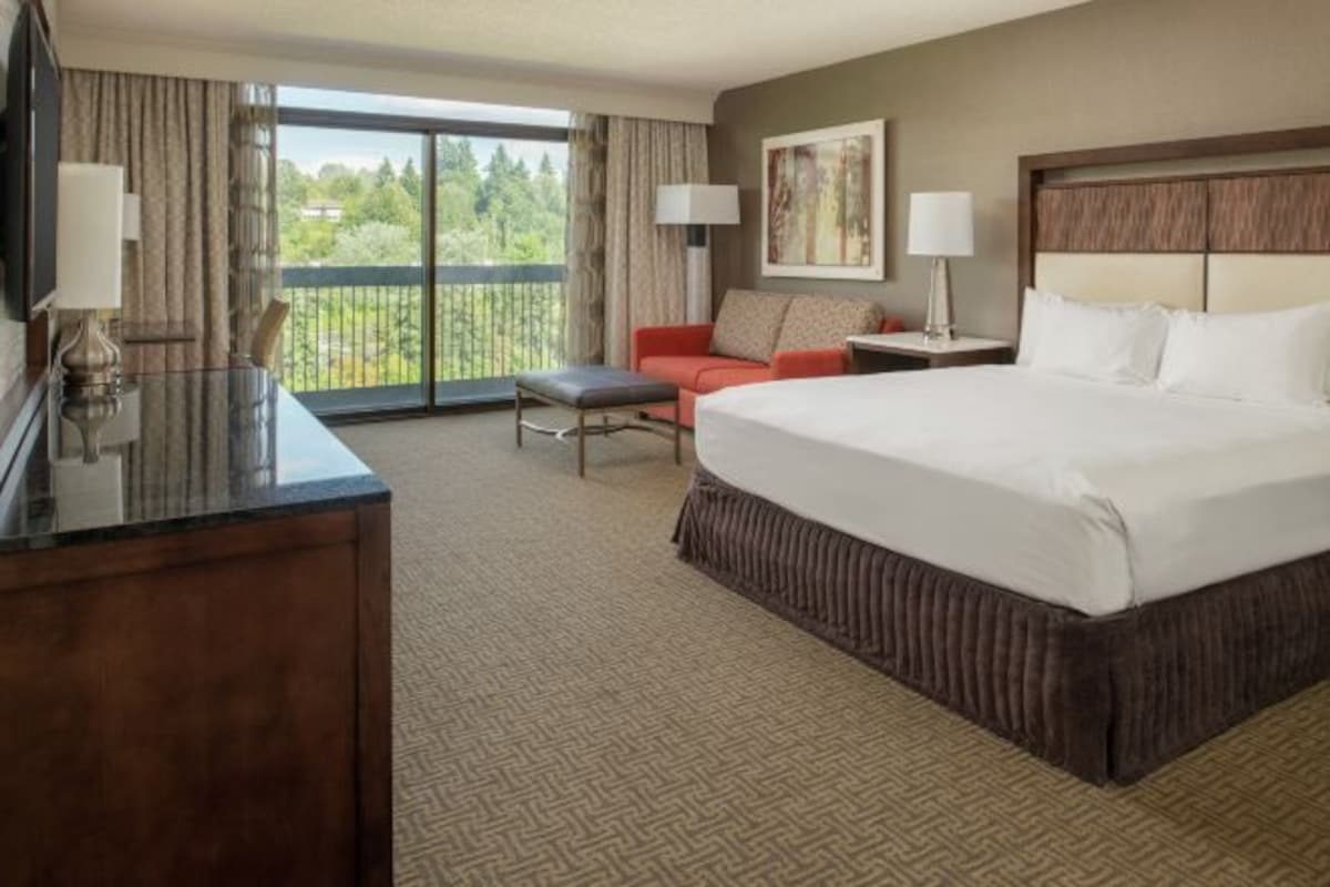 A hotel room with a large bed, a desk, a chair, a sofa, and a balcony with a view of greenery.