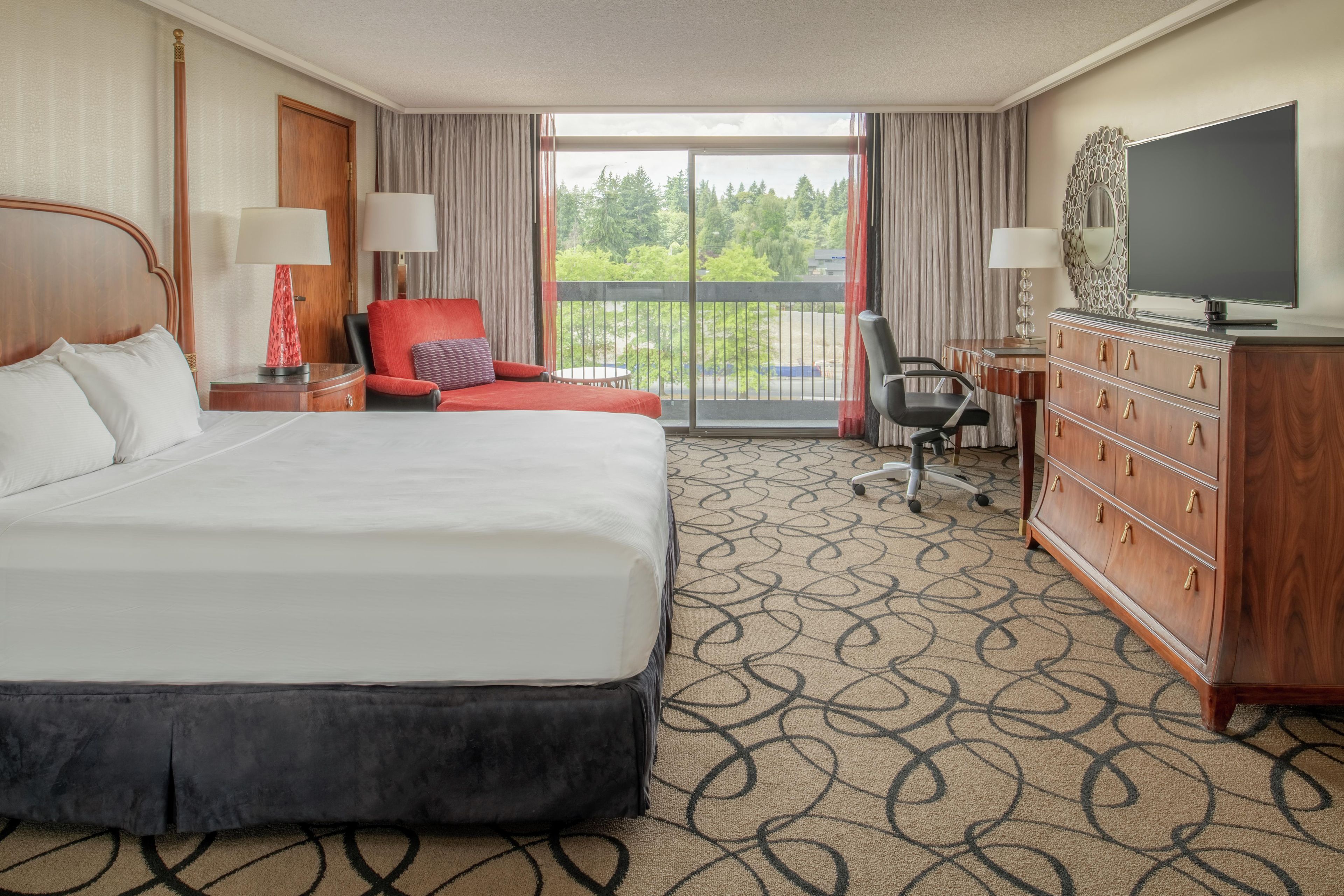 A hotel room with a large bed, a desk, a chair, a television, and a view of trees through the window.