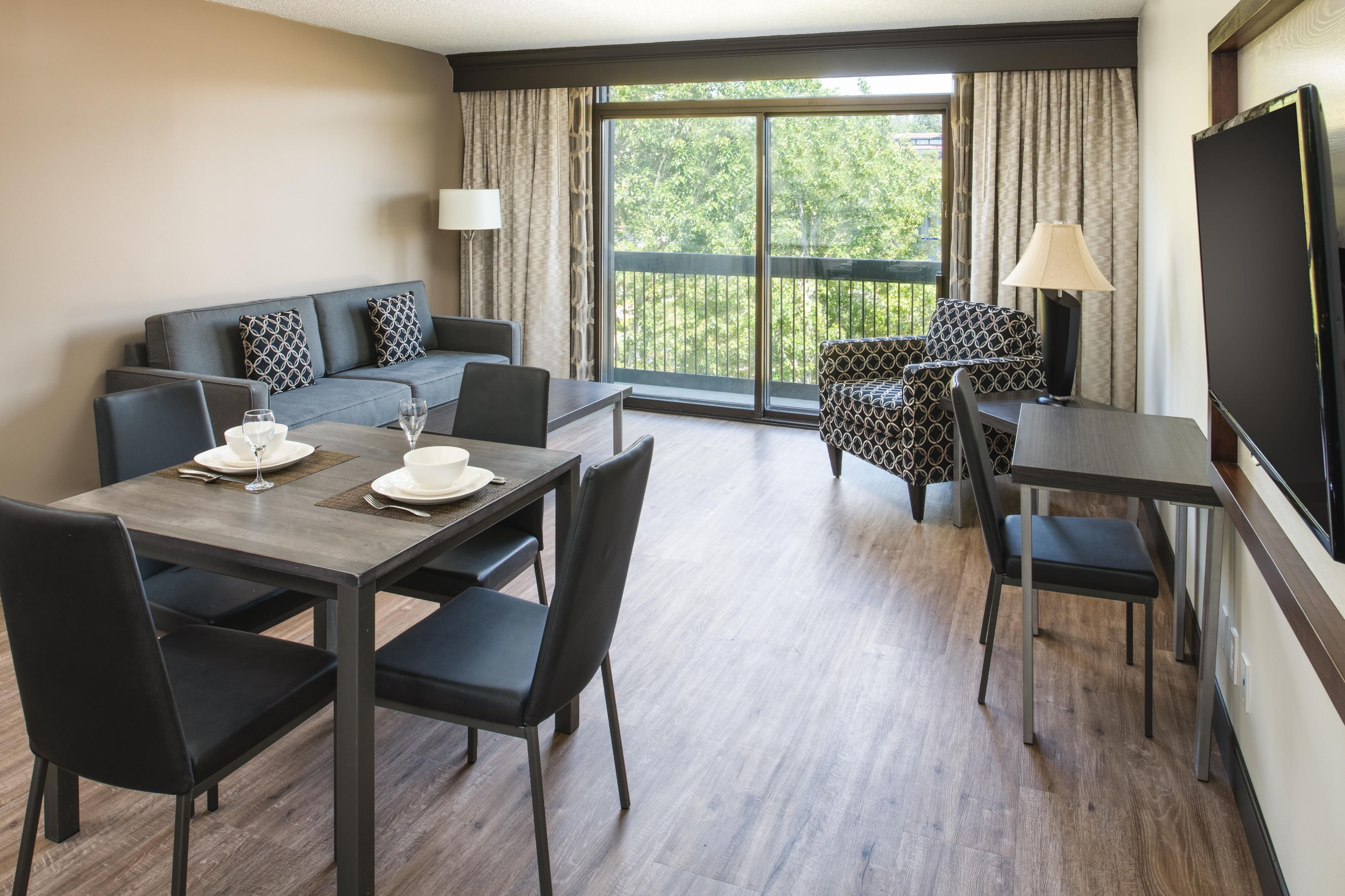 A hotel room with a dining area, a sofa, a TV, and a balcony with curtains.