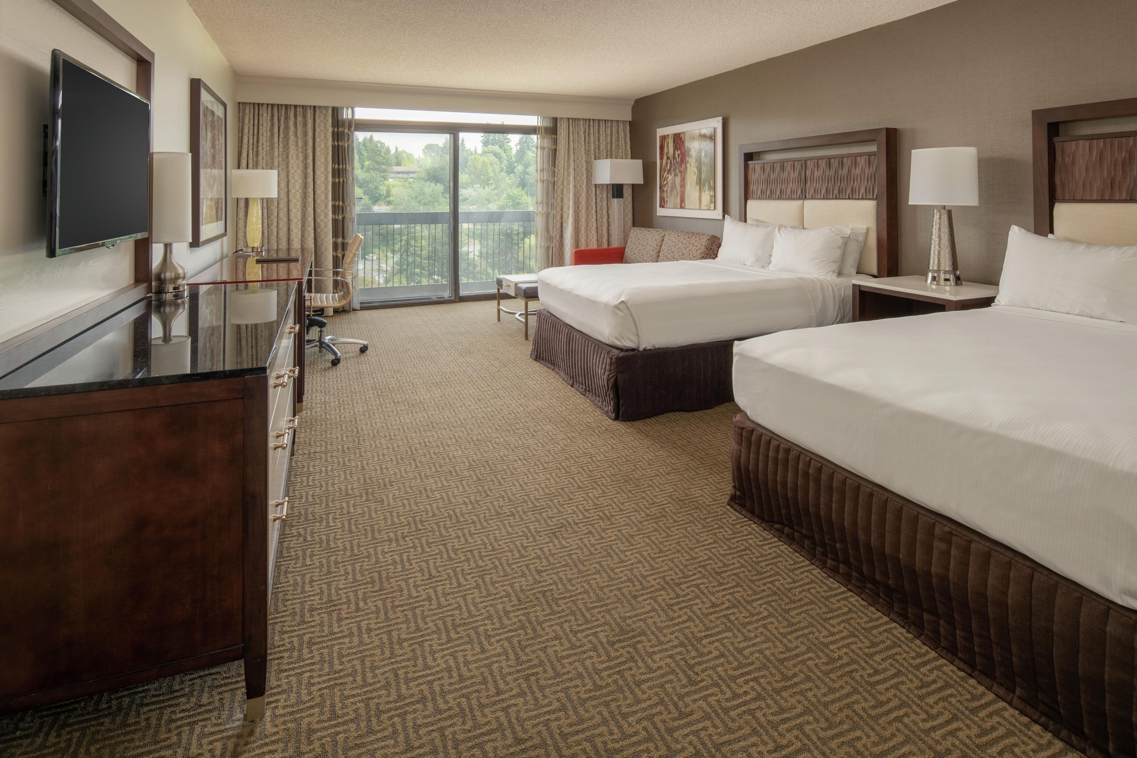 A hotel room with two beds, a desk, a TV, and a balcony with a view.