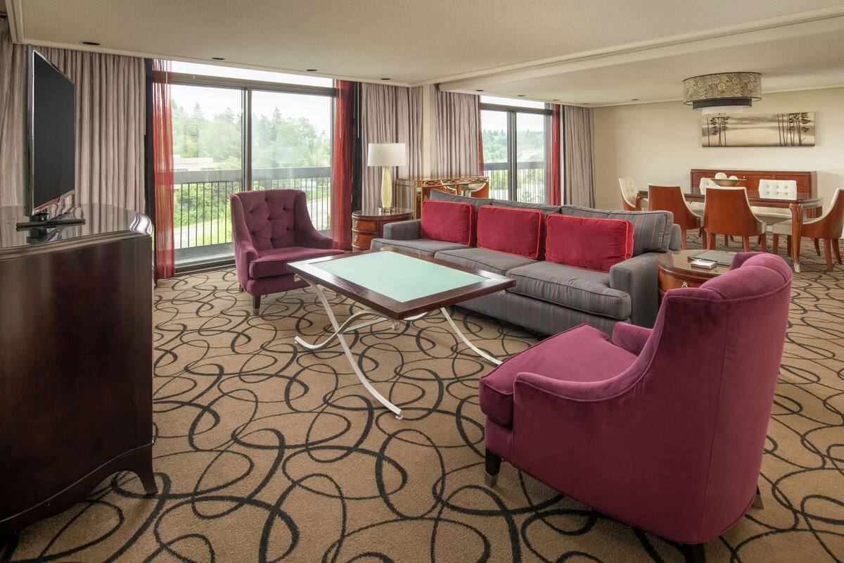 A hotel room with a large sofa, a loveseat, a coffee table, a TV, and a balcony with a dining area.