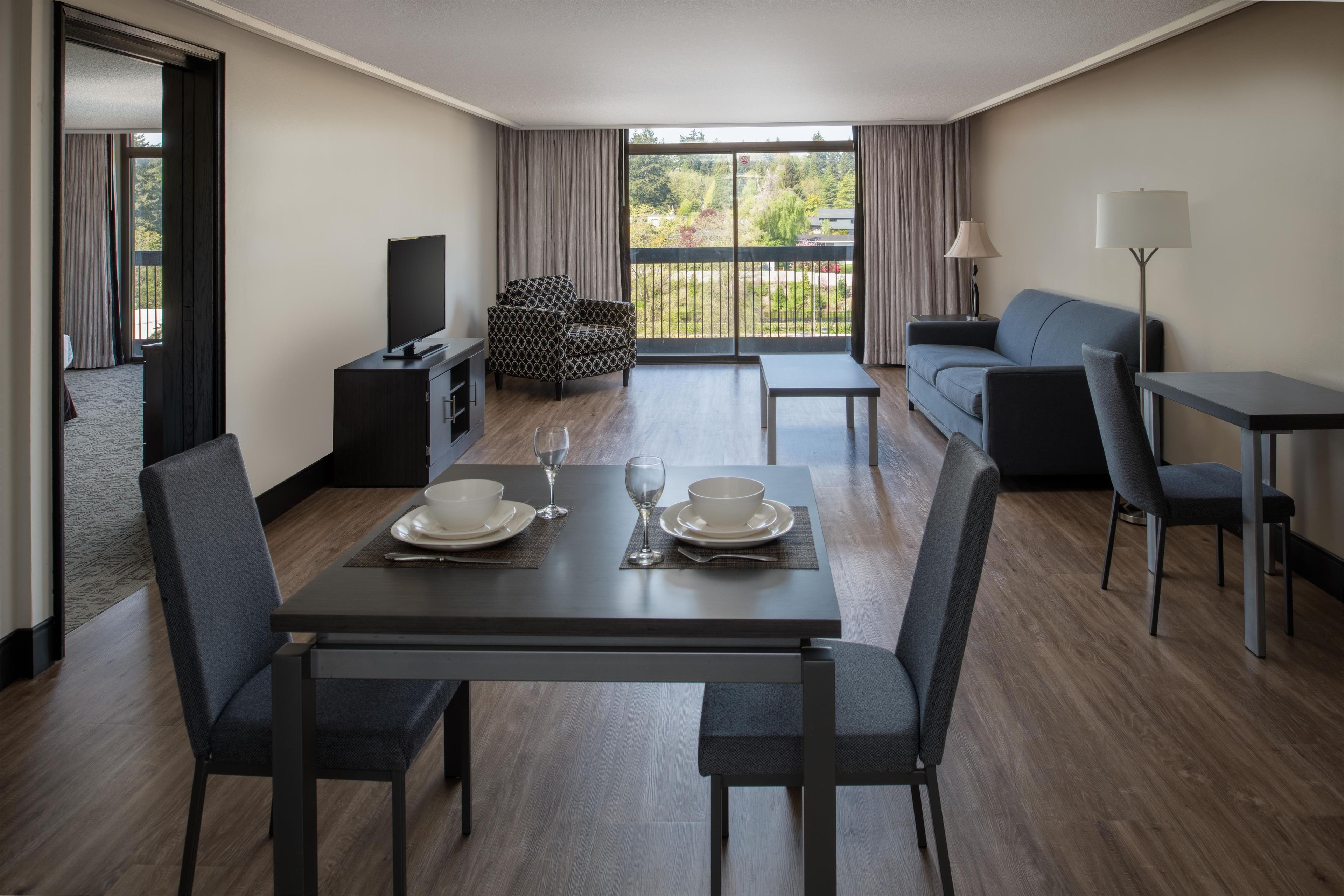 A modern hotel room with a dining table set for two, a sofa, a TV, and a balcony with a view of greenery.