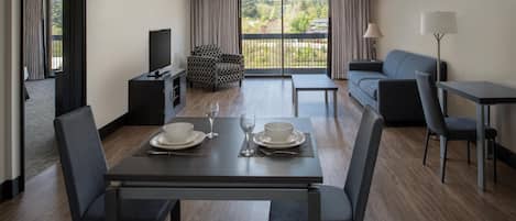Une chambre d’hôtel moderne équipée d’une table à manger pour deux, d’un canapé, d’une télévision et d’un balcon offrant une vue sur la verdure.