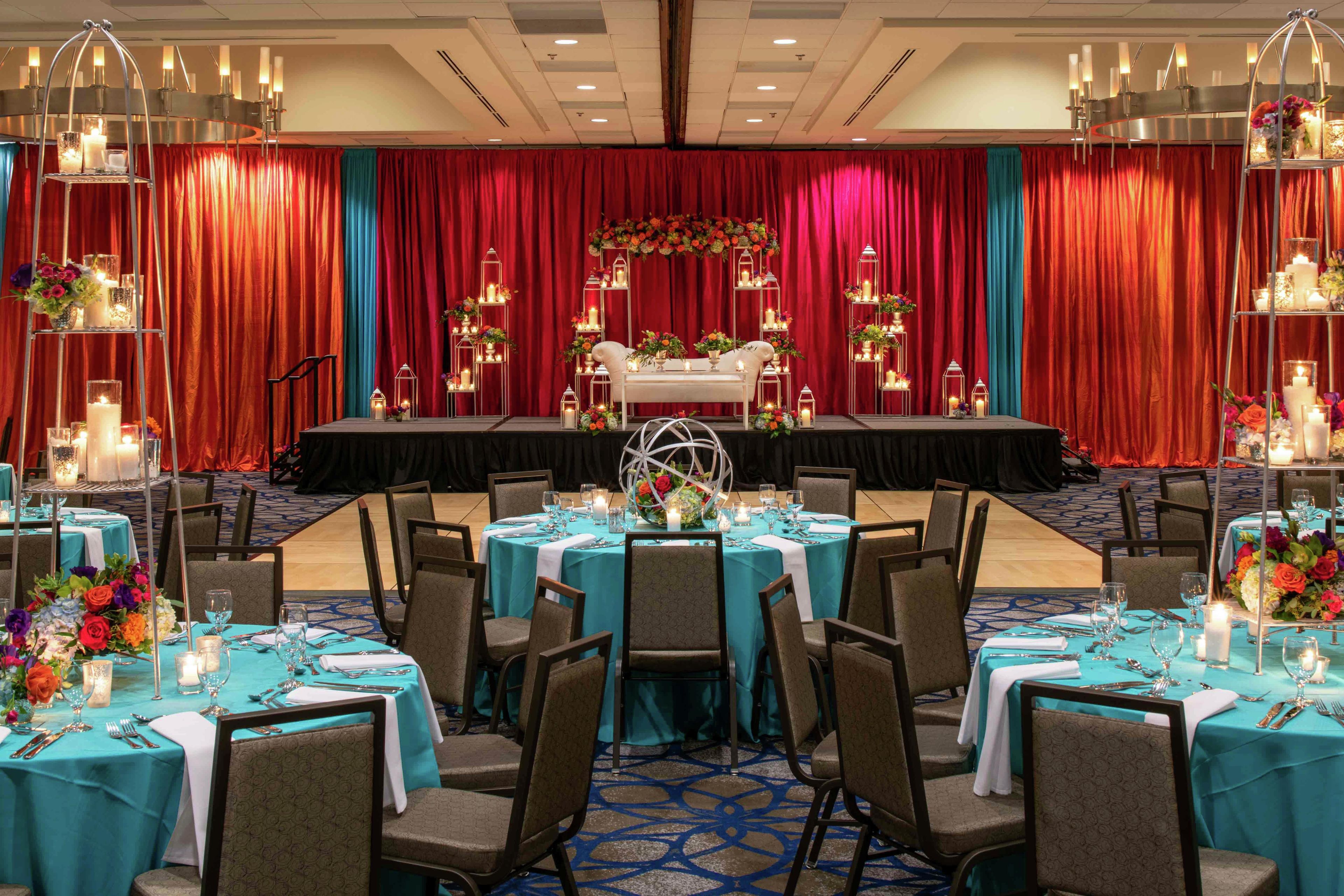 Un salón de banquetes con mesas redondas dispuestas para comer, un escenario con decoraciones florales y cortinas rojas.