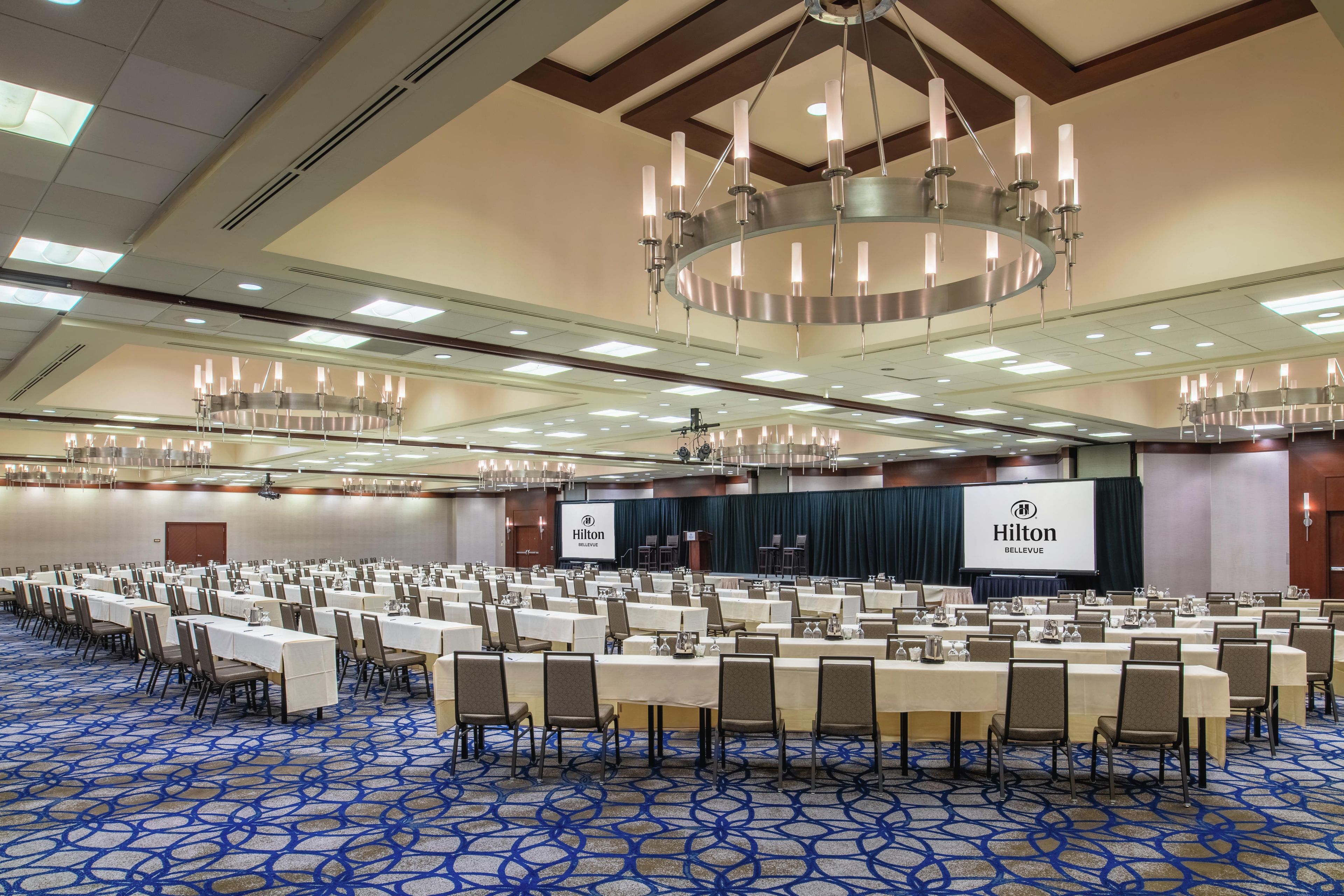 A large conference room with multiple tables and chairs, a stage with a screen, and a chandelier.
