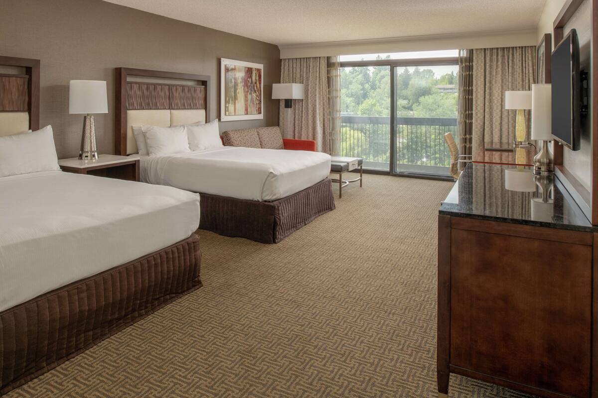 A hotel room with two beds, a desk, a TV, and a balcony with a view.