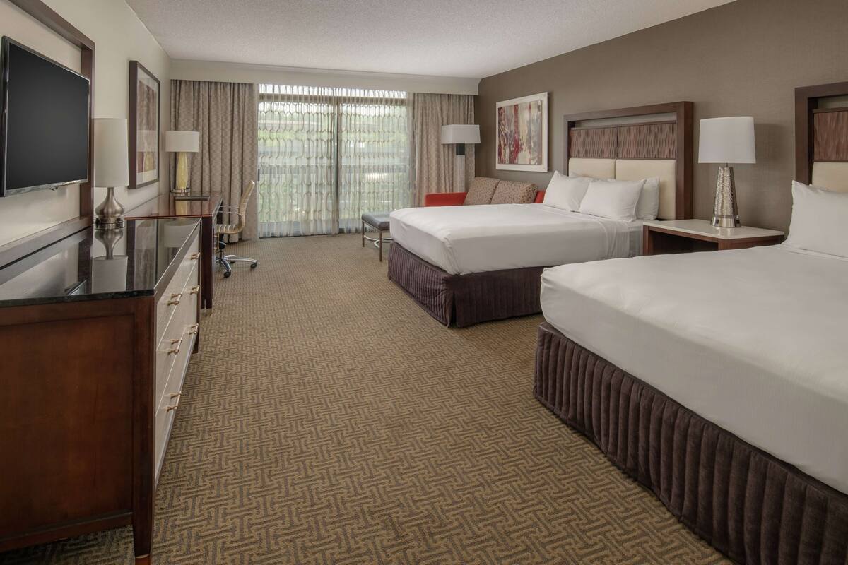 A hotel room with two beds, a desk, a TV, and a large window with curtains.
