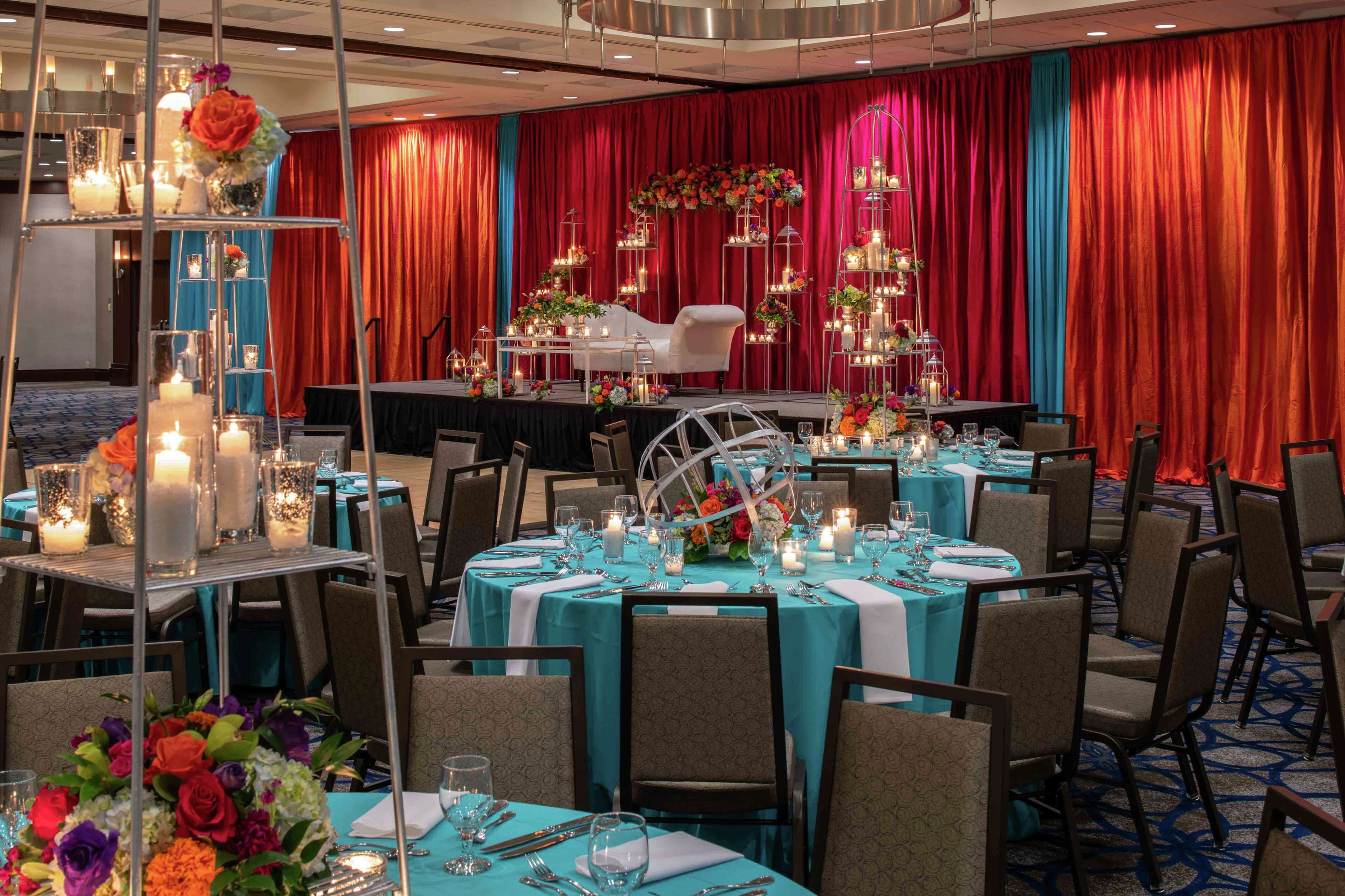 A banquet hall with round tables set for dining, a stage with floral decorations, and a red curtain backdrop.