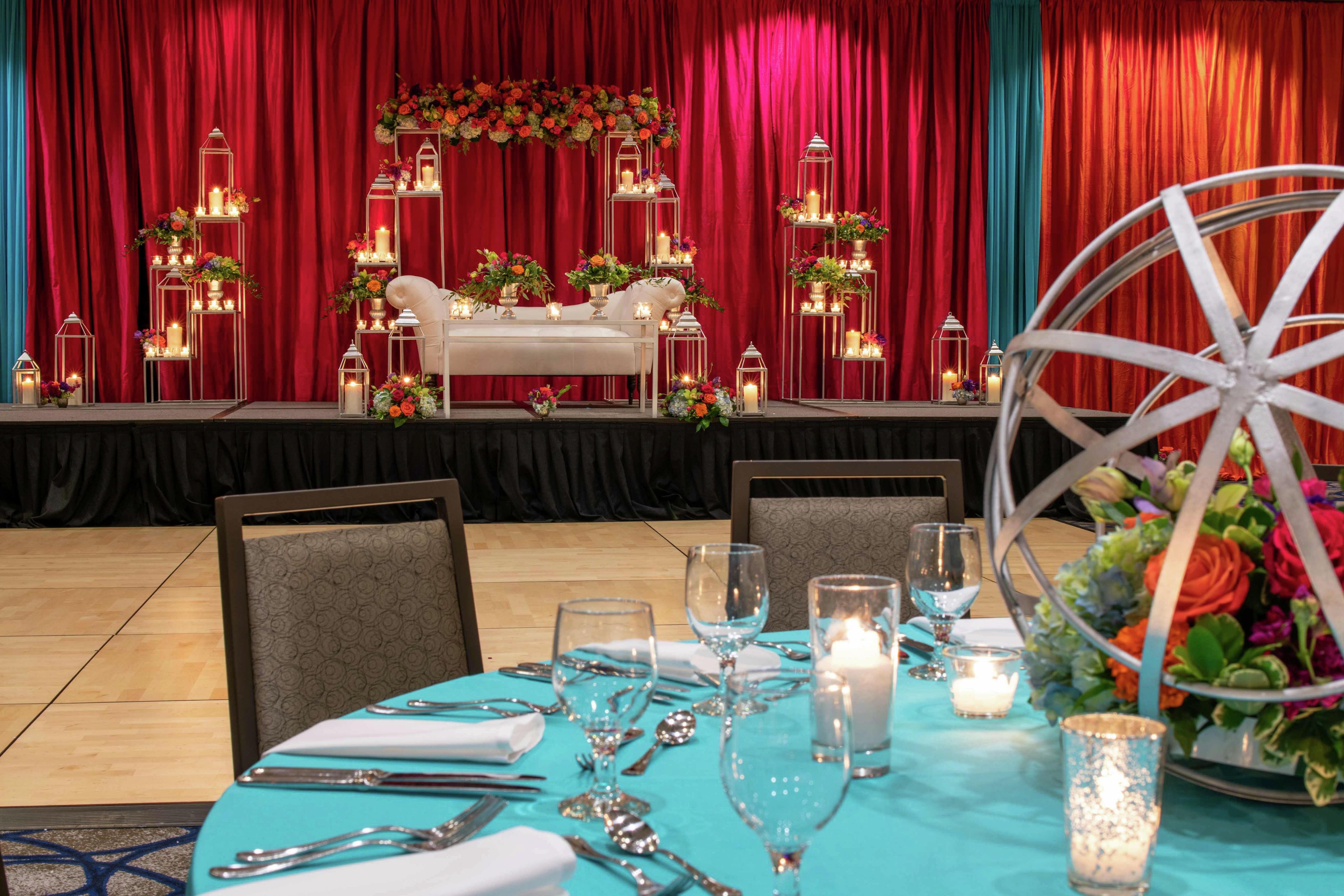 A formal dining setup with a table set for four, a stage with floral decorations, and a large wheel in the foreground.