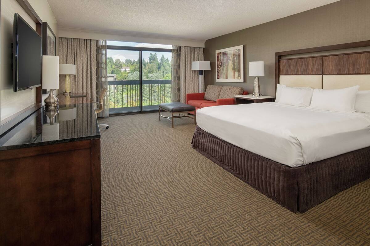 A hotel room with a large bed, a desk, a chair, a sofa, and a balcony with a view of greenery.