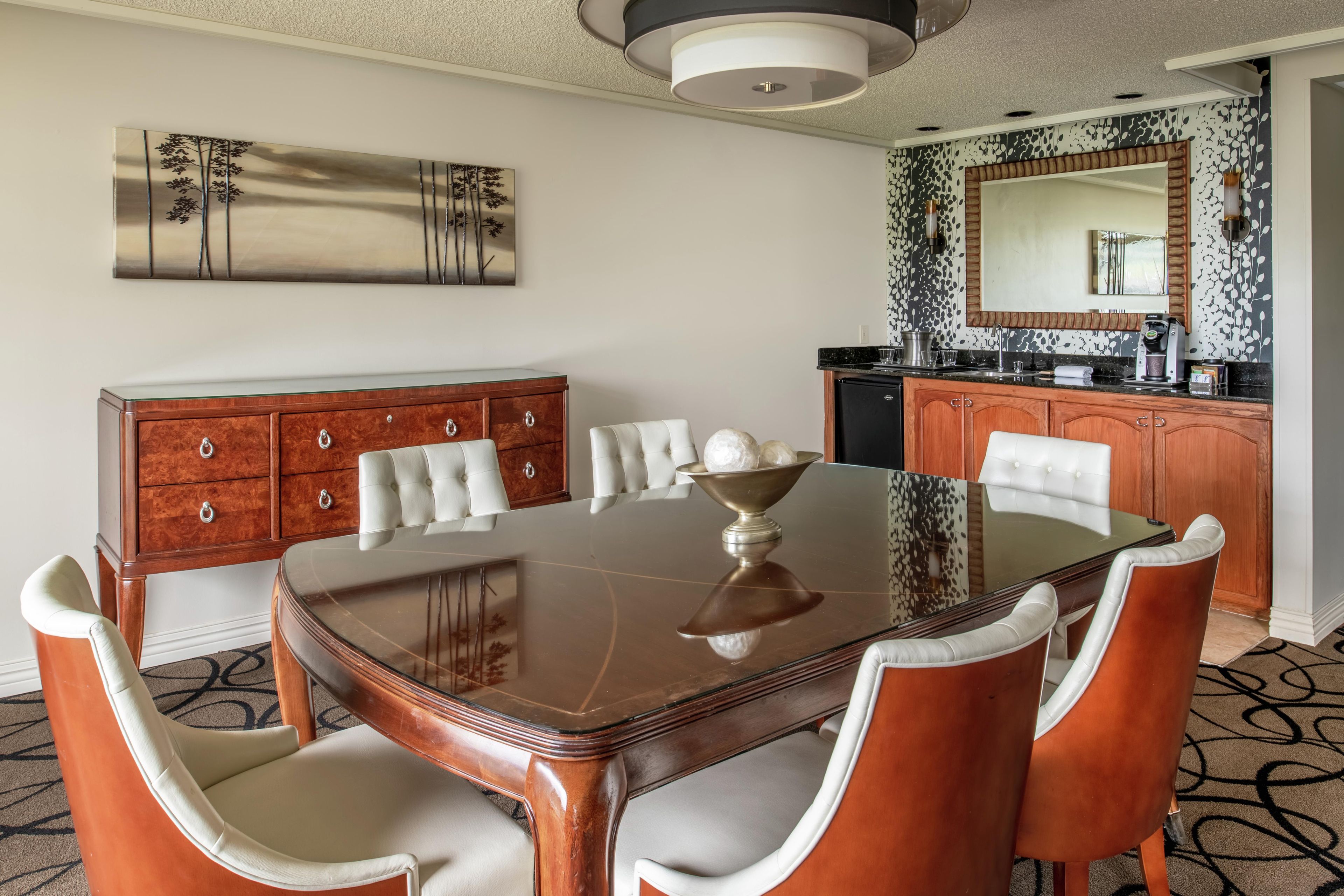 A dining area with a wooden table, white upholstered chairs, a wooden sideboard, and a large mirror above the counter.