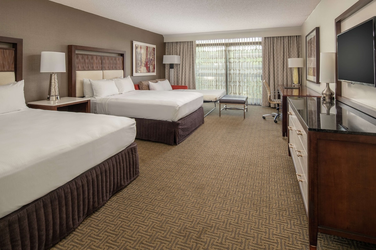 A hotel room with two beds, a desk, a TV, and a large window with curtains.