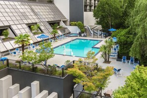 A rooftop pool area with multiple lounge chairs and umbrellas, surrounded by trees and modern architecture.