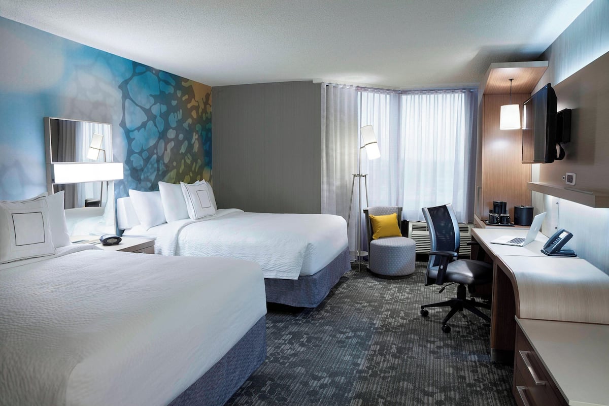 A hotel room with two beds, a desk, a chair, a TV, and a large window with curtains.
